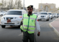 A traffic officer wearing a reflective vest and cap is directing traffic on a road in Abu Dhabi, with cars and buildings visible in the background.