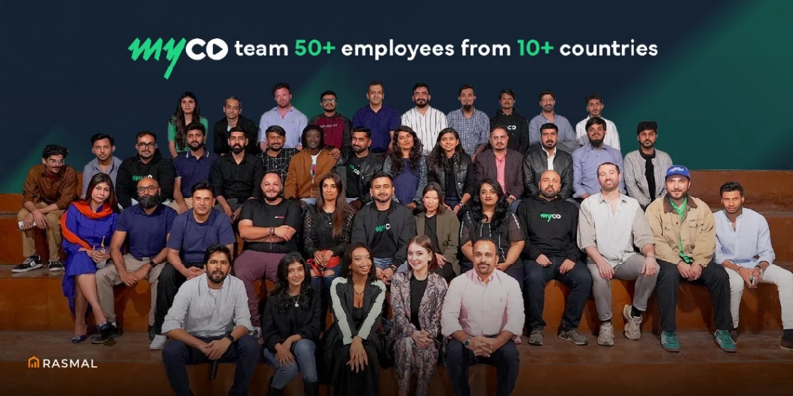 A group of people, mostly men, are smiling and looking at the camera. They are all wearing casual clothing and are sitting on a set of wooden steps. The text "myco team 50+ employees from 10+ countries" is displayed above the group.