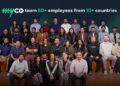 A group of people, mostly men, are smiling and looking at the camera. They are all wearing casual clothing and are sitting on a set of wooden steps. The text "myco team 50+ employees from 10+ countries" is displayed above the group.